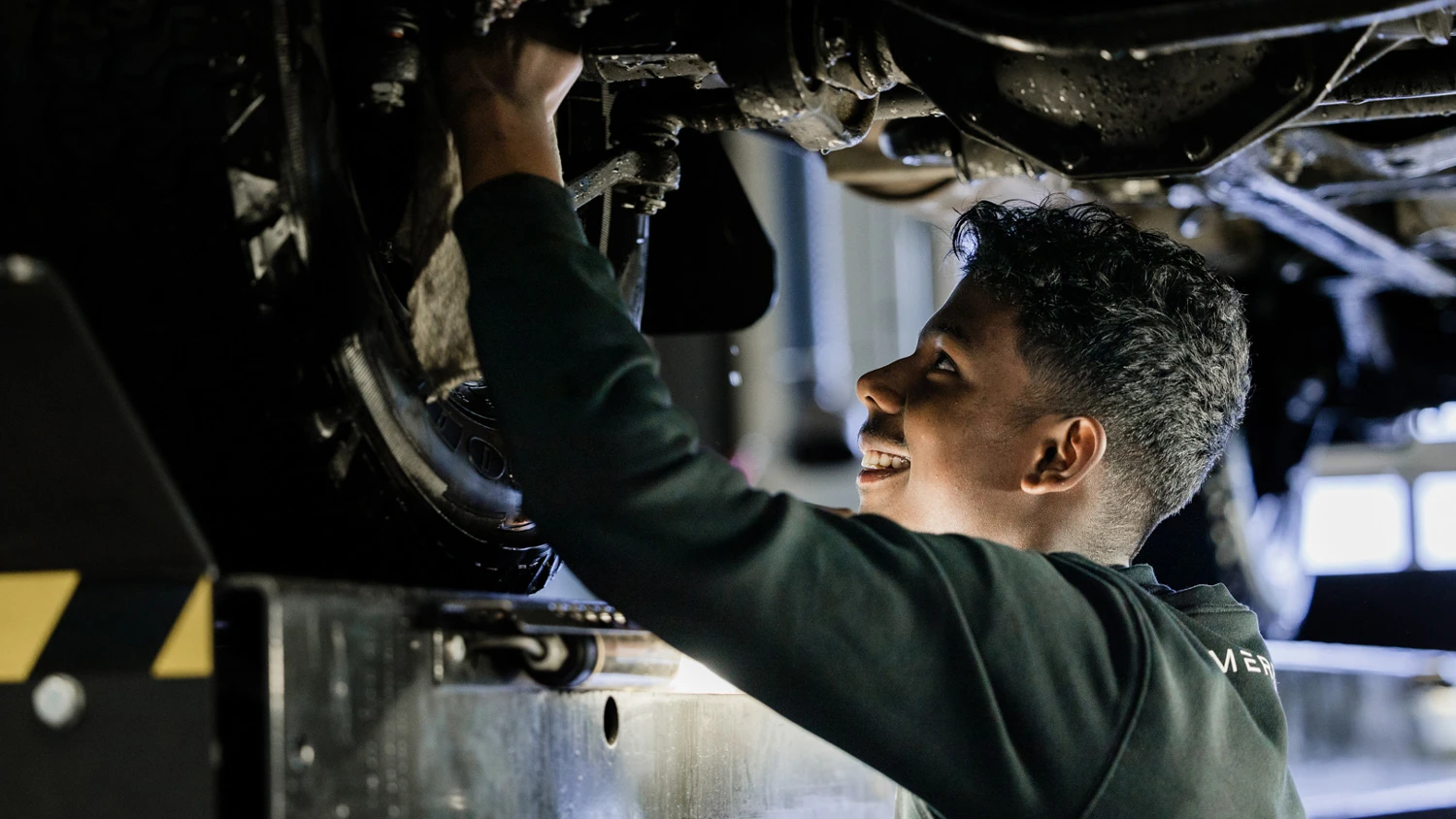 Jeune apprenti réparant le dessous d'une voiture surélevée en souriant.